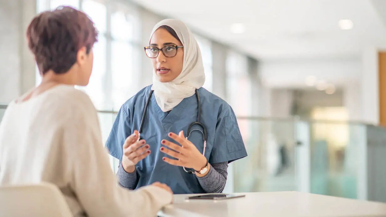 A medical professional wearing a hijab and glasses speaking to a patient. 