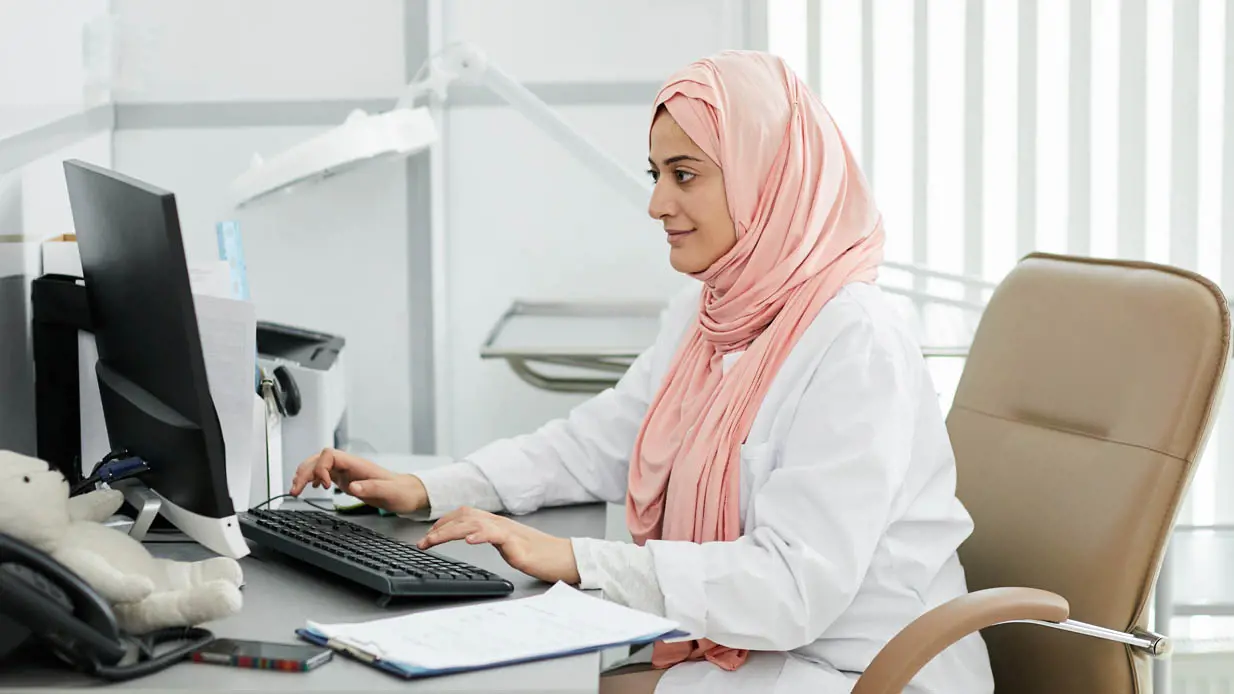 A doctor wearing a hijab working on a computer in an office. 