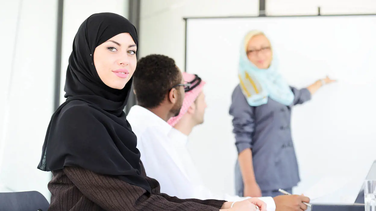 A woman wearing a hijab smiling at a camera during a meeting. 
