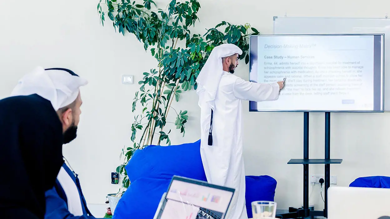 A man in a keffiyeh leading a meeting in front of a powerpoint presentation. 