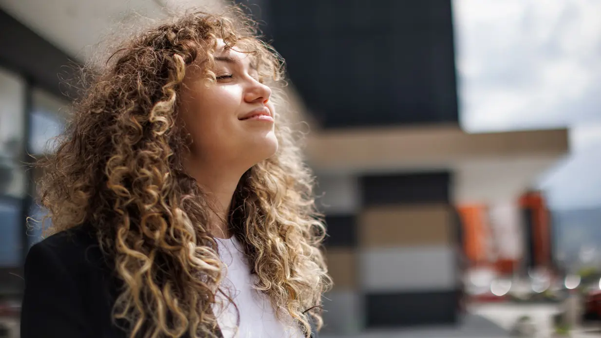 A woman with curly hair stands outdoors, surrounded by nature, exuding confidence and grace.